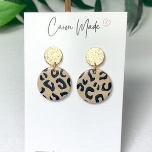 May include: Handmade earrings with a tan and black leopard print design. Each earring has a gold-coloured disc at the top and a larger, round leopard print pendant. The earrings are displayed on a white card with the text "Caron Made".