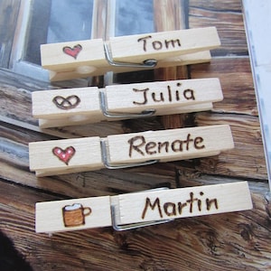 Könnte beinhalten: Vier Holz-Wäscheklammern mit eingebrannten Namen und Symbolen. Die Namen sind Tom, Julia, Renate und Martin. Die Symbole sind ein Herz, ein Unendlichkeitszeichen, ein Herz mit Punkten und ein Bierkrug.