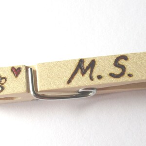 Könnte beinhalten: Ein Holz-Wäscheklammer mit den Initialen "M.S." eingebrannt in das Holz. Ein kleines Herz und eine Kaffeetasse sind ebenfalls in das Holz eingebrannt.