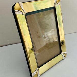May include: A rectangular picture frame with a polished gold-tone finish and silver-tone accents at each of the four corners. The frame features a black inner border surrounding a central rectangular opening, designed to hold a photograph or print.