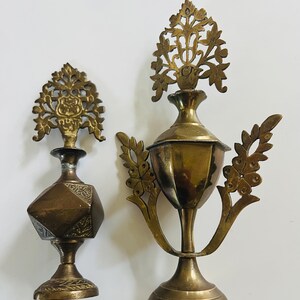 May include: Two antique brass decorative perfume bottles. The bottle on the left has a geometric design and a floral top. The bottle on the right has a cup-like shape with a floral design and leaf accents.