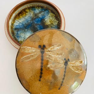 May include: A ceramic jar with a lid, featuring a dragonfly design. The lid is brown with two dragonfly illustrations. The jar's interior has a blue and gold crackle glaze. The rim is brown.