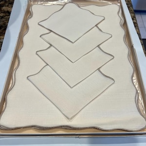 May include: A boxed set of ivory-colored linen placemats and napkins. The placemat has a scalloped edge with a gray trim, and the napkins are square with a matching gray trim. The set is neatly arranged inside a cardboard box.