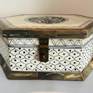 May include: An octagonal decorative box with a hinged lid. The box features a cream-colored surface with black patterned designs and a dark wood trim. A brass clasp secures the lid. The top of the box has a circular black and white design.
