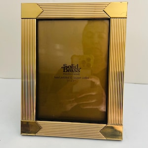 May include: A rectangular solid brass photo frame with a polished gold finish and ribbed texture. The frame features smooth corner accents and a central insert displaying the text Solid Brass PHOTO FRAME hand polished & lacquer coated 5" x 7".