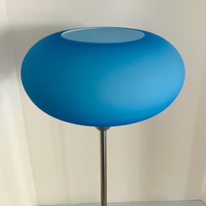 May include: A floor lamp featuring a large, rounded, frosted blue glass shade with a smooth, matte texture. The shade is shaped like a flattened sphere with an open top. It is supported by a slender, straight, metallic pole with a neutral finish.