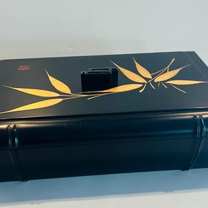 May include: A rectangular black box with a smooth finish, featuring a gold-colored bamboo leaf design on the lid. The lid includes a small black handle and a red square stamp mark. The box has rounded edges and a sleek, dark appearance.