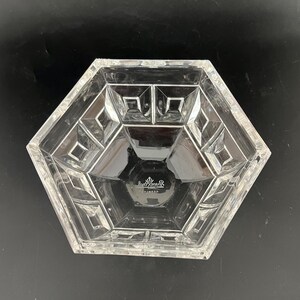 May include: Clear, hexagonal glass bowl with geometric designs. The bowl has a faceted exterior and a smooth interior. The base of the bowl has a logo. A decorative piece for serving or display.