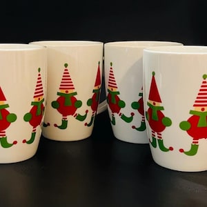 May include: Set of four white ceramic mugs with a Christmas elf design. Each mug features a red elf with a green scarf and white hat, standing on green and white striped skis.