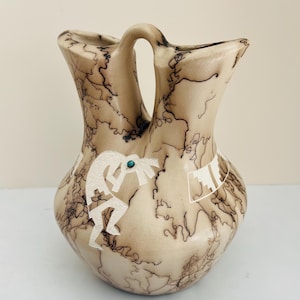 May include: A tan-colored ceramic vase with a unique double-spout design and a handle. The vase features a white Kokopelli figure and geometric patterns, accented with a turquoise stone. The surface is decorated with dark, lightning-like patterns.