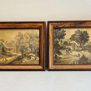 May include: Two rectangular wall art pieces in dark brown wood frames. The left print depicts a house with trees and grazing sheep. The right print shows a water mill, a stone bridge, and a house near a road. Both feature sepia-toned landscapes.