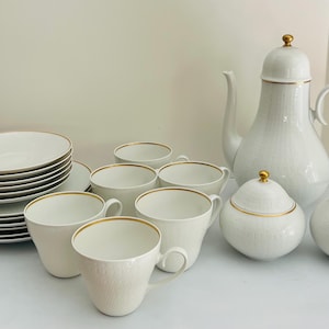 May include: A white porcelain tea set with gold trim. The set includes a teapot, sugar bowl, creamer, cups, saucers, and plates. The teapot has a tall, slender design with a lid. The cups and plates are stacked.