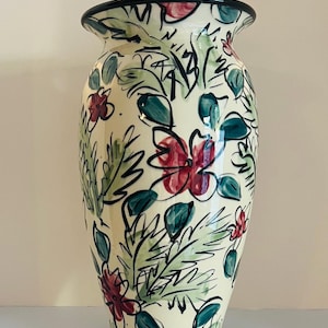 May include: A tall, cream-colored ceramic vase with a black rim and a flared top. The vase is adorned with a hand-painted floral design featuring red flowers, green leaves, and black outlines. The vase is approximately 12 inches tall.