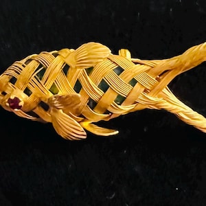 May include: A gold-toned fish brooch with a woven basket design. The fish has detailed fins and tail, with a small red gemstone eye. The brooch is set against a dark background, highlighting the intricate metalwork.