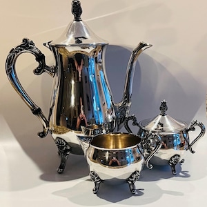 May include: A silver tea set with ornate detailing. The set includes a tall coffee pot, a creamer, a sugar bowl with a lid, and a small pitcher. The pieces have decorative handles and are set on small, decorative feet.