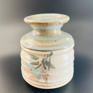 May include: A small, white ceramic vase with a blue and brown glaze. The vase has a simple, rounded shape and a wide mouth. It is decorated with a hand-painted design of stylized leaves in shades of blue, green, and pink.