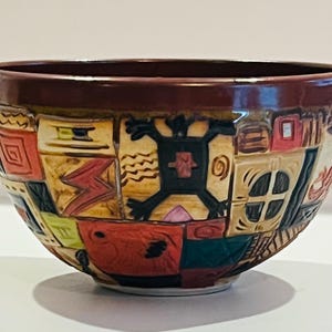 May include: A ceramic bowl with a patchwork design featuring geometric shapes, trees, and houses in various colors. The rim is a dark reddish-brown, and the bowl has a glossy finish. The design is carved into the surface.