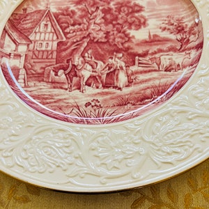 May include: A vintage ceramic plate with a raised rim and a detailed floral design. The center of the plate features a red and white illustration of a rural scene with a farmhouse, trees, and people.