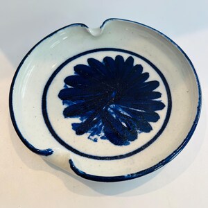May include: Round ceramic ashtray with a white base and a blue floral design in the center. The ashtray has a dark blue rim and a notch for resting cigarettes. The floral design is a deep shade of blue.
