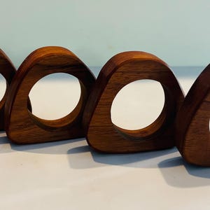 May include: A set of four brown wooden napkin rings, each featuring a rounded triangular shape with a circular opening in the center. The wood grain is visible on the smooth, polished surfaces of these four individual dining table accessories.