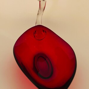 May include: A red glass dish with a clear glass handle. The dish is shaped like a scoop and has a rounded bottom.