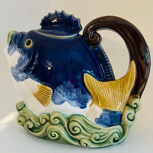 May include: A blue and white ceramic teapot shaped like a fish with a brown handle. The fish is swimming over green waves.