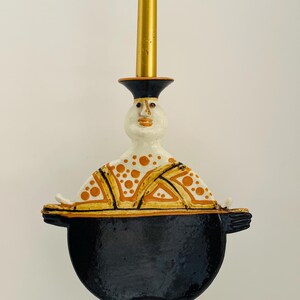 May include: A ceramic candle holder shaped like a stylized figure with a white head and torso featuring orange polka dots and geometric lines. The figure has a black base and a large black bowl-like midsection, topped with a tall gold taper candle.