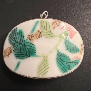 May include: Oval shaped pendant with a white background and a floral design. The design features green leaves and pink flowers.