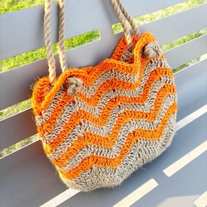 Könnte beinhalten: Handgefertigte Häkeltasche mit Zickzackmuster in Orange und Grau. Die Tasche hat Seilgriffe und einen Kordelzugverschluss. Die Tasche liegt auf einer grauen Bank.