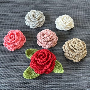 Può includere: Una collezione di sei fiori all'uncinetto fatti a mano in vari colori. I fiori includono sfumature di rosa, rosso, beige, grigio e bianco. Un fiore rosso ha foglie verdi. I fiori sono disposti su una superficie grigia.