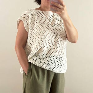 May include: Cream-colored crochet top with a diamond pattern. The top has short sleeves and a scoop neckline. The person in the image is wearing olive green pants. The top is handmade and has a loose, airy design.