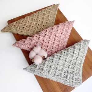 May include: Three crocheted triangle-shaped cloths in beige, pink, and gray on a wooden surface. The cloths are made with a waffle stitch pattern.