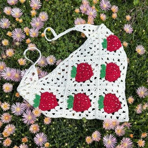 Könnte beinhalten: Eine weiße Häkeltasche mit Erdbeermuster. Die Tasche hat rote Erdbeeren mit grünen Blättern auf einem weißen, offen gewebten Hintergrund. Die Tasche hat einen Griff zum Tragen.