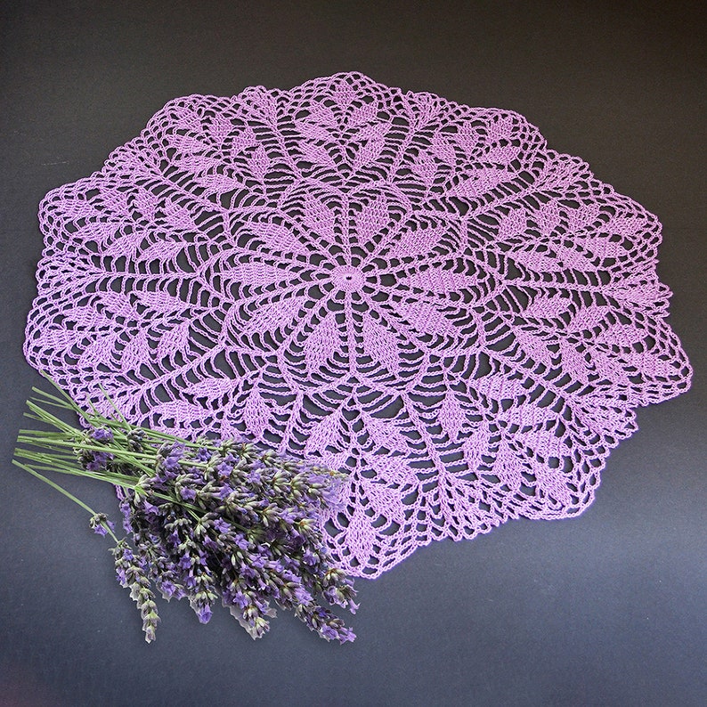 Puede incluir: Un delicado encaje de ganchillo de color morado con un dise&ntilde;o circular, que presenta un motivo central y un trabajo de encaje intrincado. Un ramo de flores de lavanda se coloca sobre el encaje.