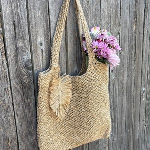 May include: A beige crocheted tote bag with a fringe detail hanging on a wooden wall. The bag has a single strap and a feather-shaped embellishment.