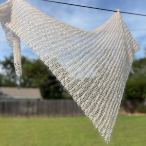 Può includere: Uno scialle triangolare beige chiaro realizzato in un materiale a maglia aperta. Lo scialle è appeso a un filo all'aperto, con uno sfondo sfocato di erba verde e alberi sotto un cielo blu.