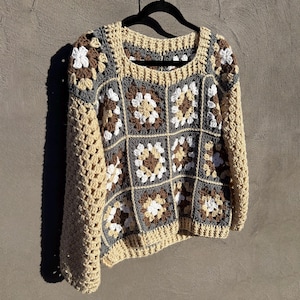 Op de afbeelding: Een gebreide trui met een granny square patroon in tinten beige, bruin, wit en grijs. De trui heeft lange mouwen en een ronde hals.