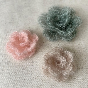 Può includere: Tre decorazioni floreali fatte a mano, soffici, in morbidi colori pastello. Uno è rosa chiaro, un altro è verde salvia tenue e il terzo è un beige chiaro. I fiori sono fatti di un materiale morbido e strutturato, possibilmente filato o fibra.