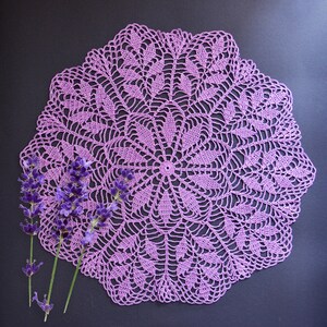 Puede incluir: Un encaje de ganchillo morado con un delicado patr&oacute;n floral. El encaje est&aacute; colocado plano sobre una superficie negra y rodeado de ramitas de lavanda.