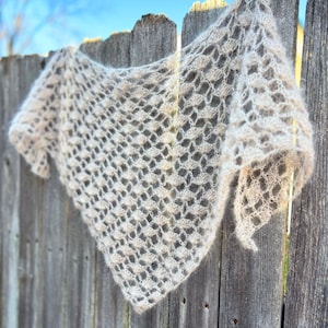May include: A light beige, open-weave shawl is draped over a weathered wooden fence. The shawl has a delicate, lacy pattern and a soft, fuzzy texture. The background shows a clear blue sky.