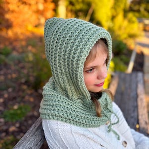 Könnte beinhalten: Eine hellgrüne gehäkelte Kapuze mit Kordelzug. Die Kapuze ist aus strukturiertem Garn gefertigt und wird über einem weißen Pullover getragen. Der Hintergrund ist verschwommen, mit Herbstfarben.