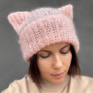 Può includere: Un cappello rosa chiaro lavorato a maglia con orecchie da gatto. Il cappello è realizzato in un materiale morbido e soffice e ha una trama a coste. Il cappello è di colore rosa chiaro e si indossa sulla testa. Un accessorio carino ed elegante.