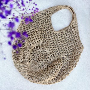 May include: A beige, open-weave tote bag with a single handle. The bag is made of a natural fiber, possibly jute or hemp, and has a textured appearance. The bag is empty and sits on a white surface, with purple flowers in the background.