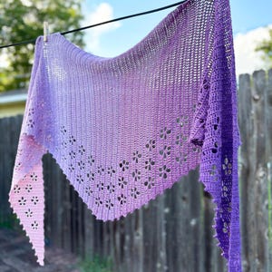 Peut inclure: Un châle crocheté à la main, dans des tons de violet et de lavande, suspendu à un fil à linge. Le châle présente un motif floral sur les bords et un effet de couleur dégradé. Le châle est fait de fil doux.