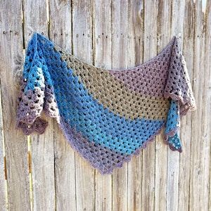 May include: A handmade crochet shawl in shades of blue, purple, and brown, hanging against a wooden fence. The shawl features a triangular shape with a scalloped edge and a gradient colour pattern, showcasing a blend of soft, muted tones.