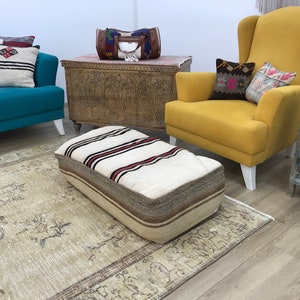 May include: A white and brown striped ottoman with a red stripe sits on a beige rug in a living room. A yellow armchair with white legs and a teal sofa with white legs are in the background.