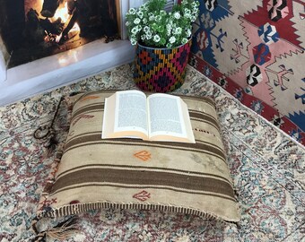 May include: A brown and beige striped floor cushion with tassels sits on a patterned rug with a book on top. The cushion has a fringe border and is made of woven fabric.