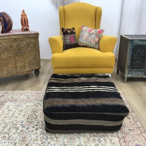 May include: A yellow armchair with two patterned throw pillows sits in a room with a wooden chest, a small wooden cabinet, and a black and white striped ottoman. The room has a light wood floor and a patterned rug.