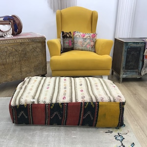 Pode incluir: Um otomano estampado colorido com um design amarelo, vermelho, preto e branco. O otomano está sentado em um tapete branco na frente de uma poltrona amarela com duas almofadas estampadas.