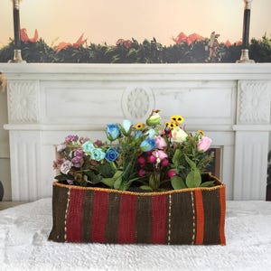 Puede incluir: Una cesta rectangular tejida con rayas verticales en marrón oscuro, rojo intenso y naranja. La cesta está llena de un surtido de flores artificiales en tonos azul, rosa, amarillo y morado, con hojas y tallos de color verde.